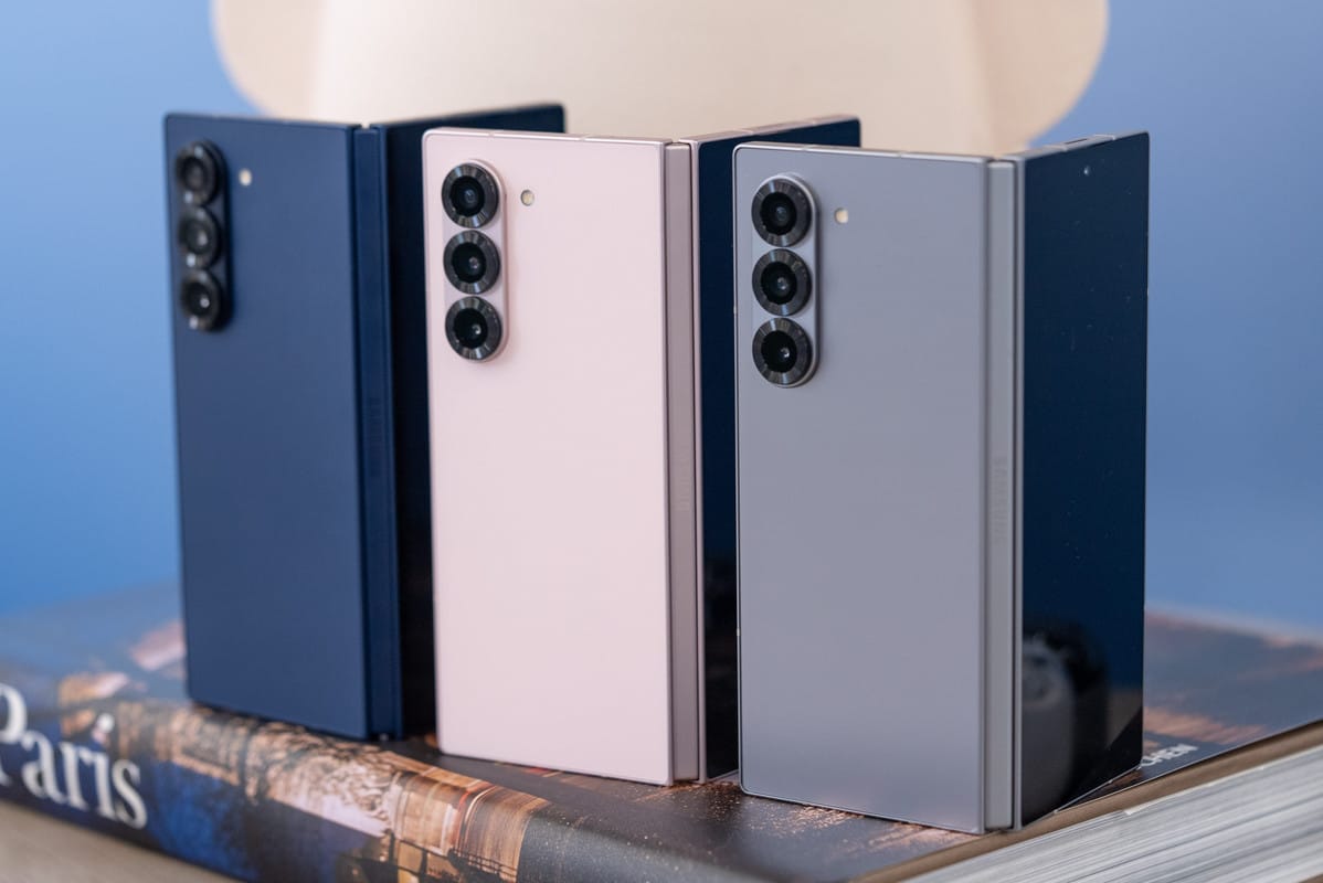 Samsung 摺疊手機 多色機身展示，藍色、粉色與灰色款直立排列，背面三鏡頭模組與機身摺疊結構呈現，置於書本上拍攝
