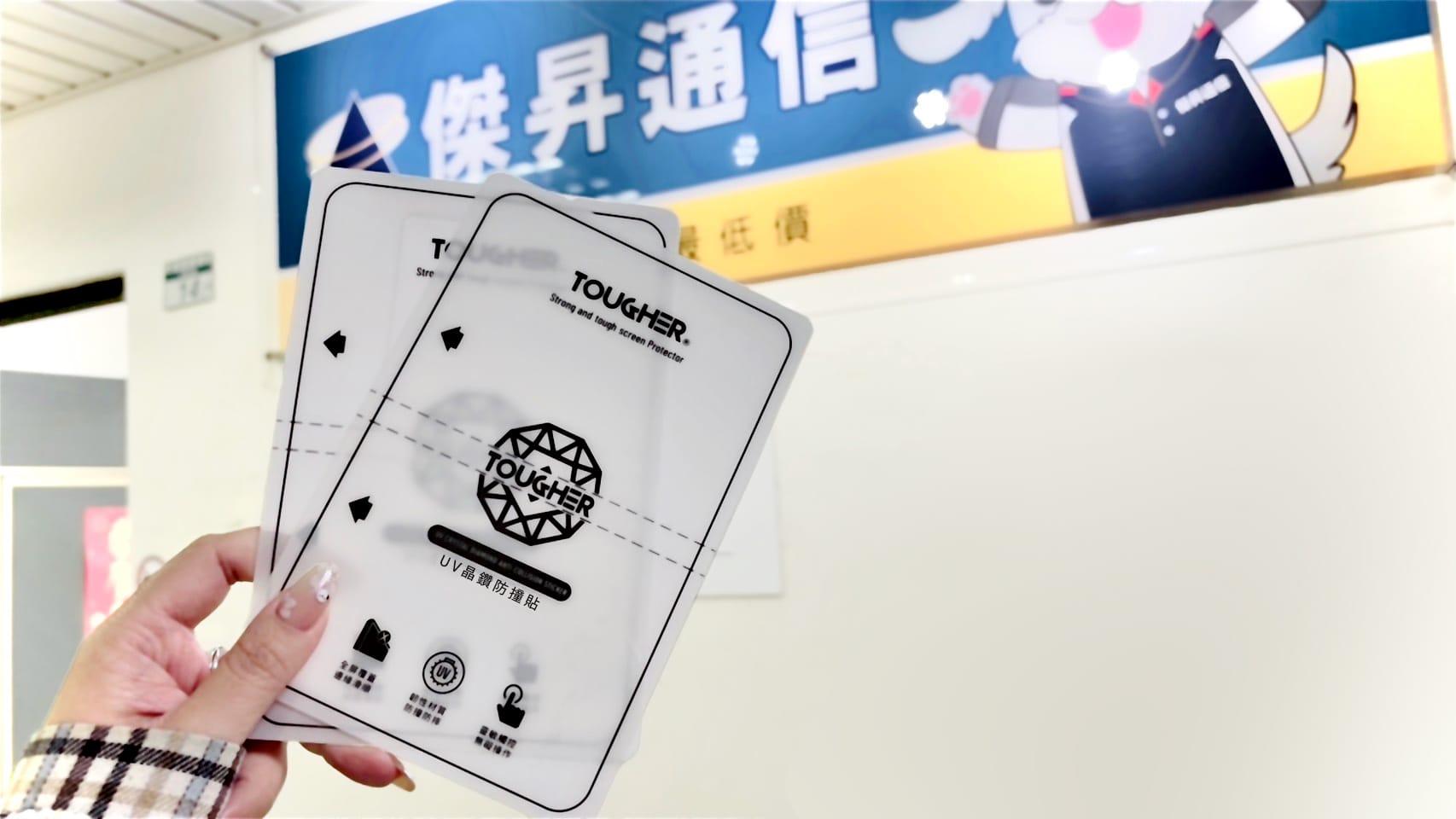 手持兩片 TOUGHER「UV晶鑽防撞貼」保護貼，背景為傑昇通信的藍色招牌看板。