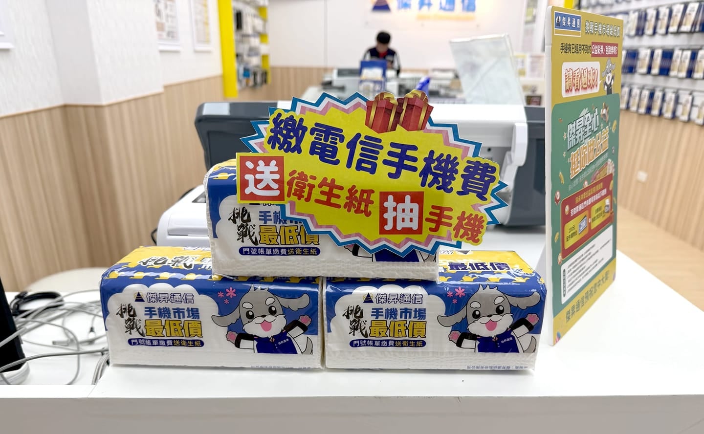傑昇通信門市櫃檯特寫,展示「繳電信手機費送衛生紙抽手機」活動立牌與專屬包裝衛生紙贈品。